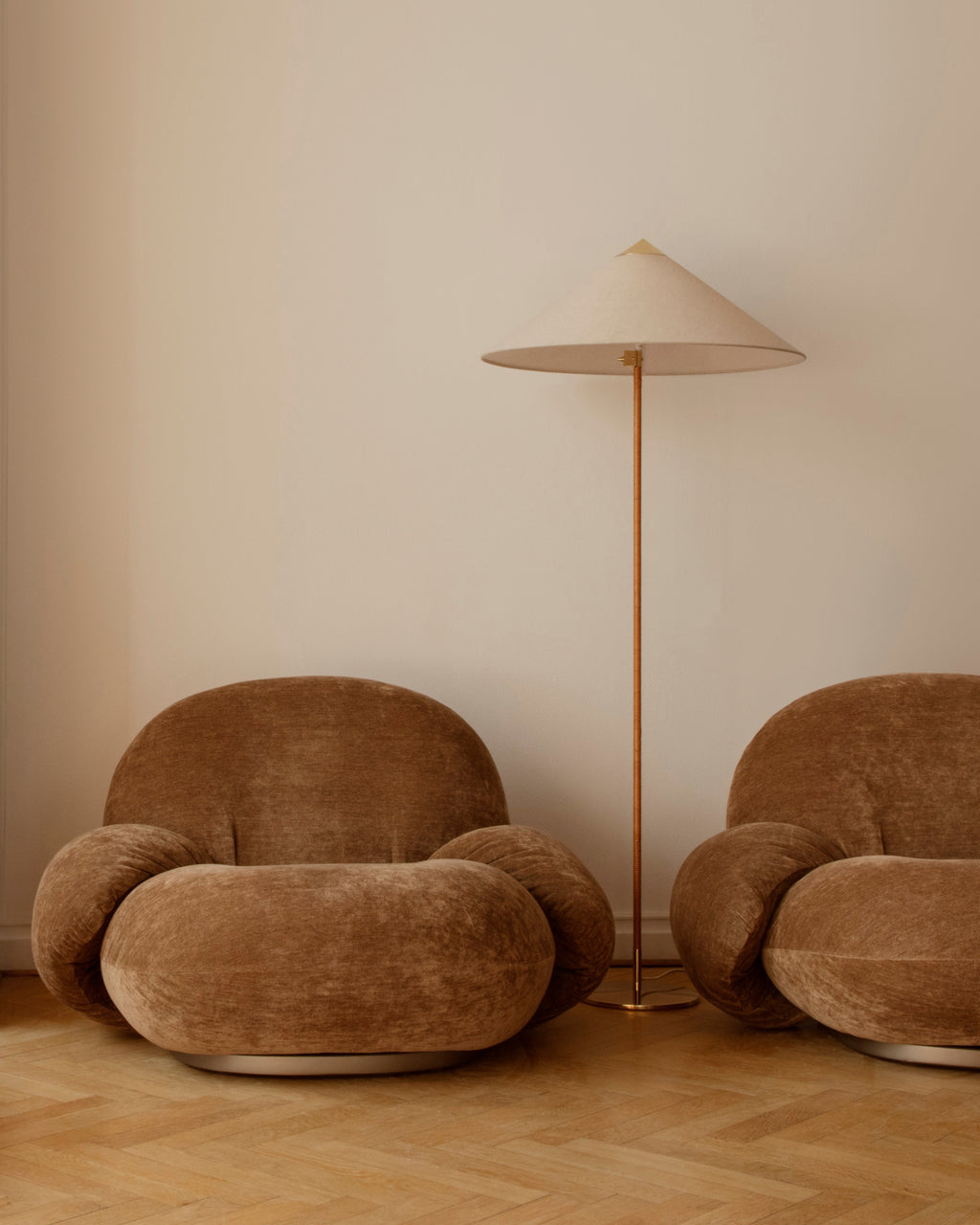 Shown here, a Clérachic floor lamp with a cream shade stands in front of a warm beige wall, between two chunky brown velvet armchairs.
