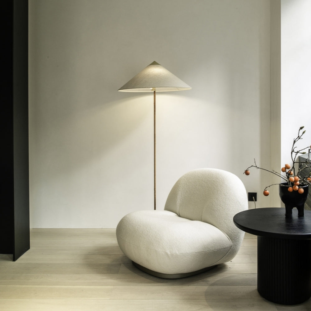 This Clérachic floor lamp with a pale shade stands lit in a quiet room, beside a rounded white armchair and a dark side table.