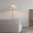 A Clérachic floor lamp with a pink-tinted white shade glows above the back of a grey sofa, against a pale wall.