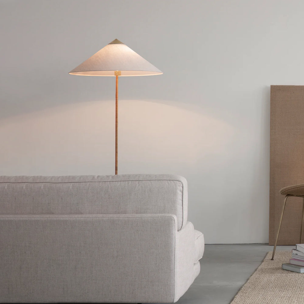 A Clérachic floor lamp with a pink-tinted white shade glows above the back of a grey sofa, against a pale wall.