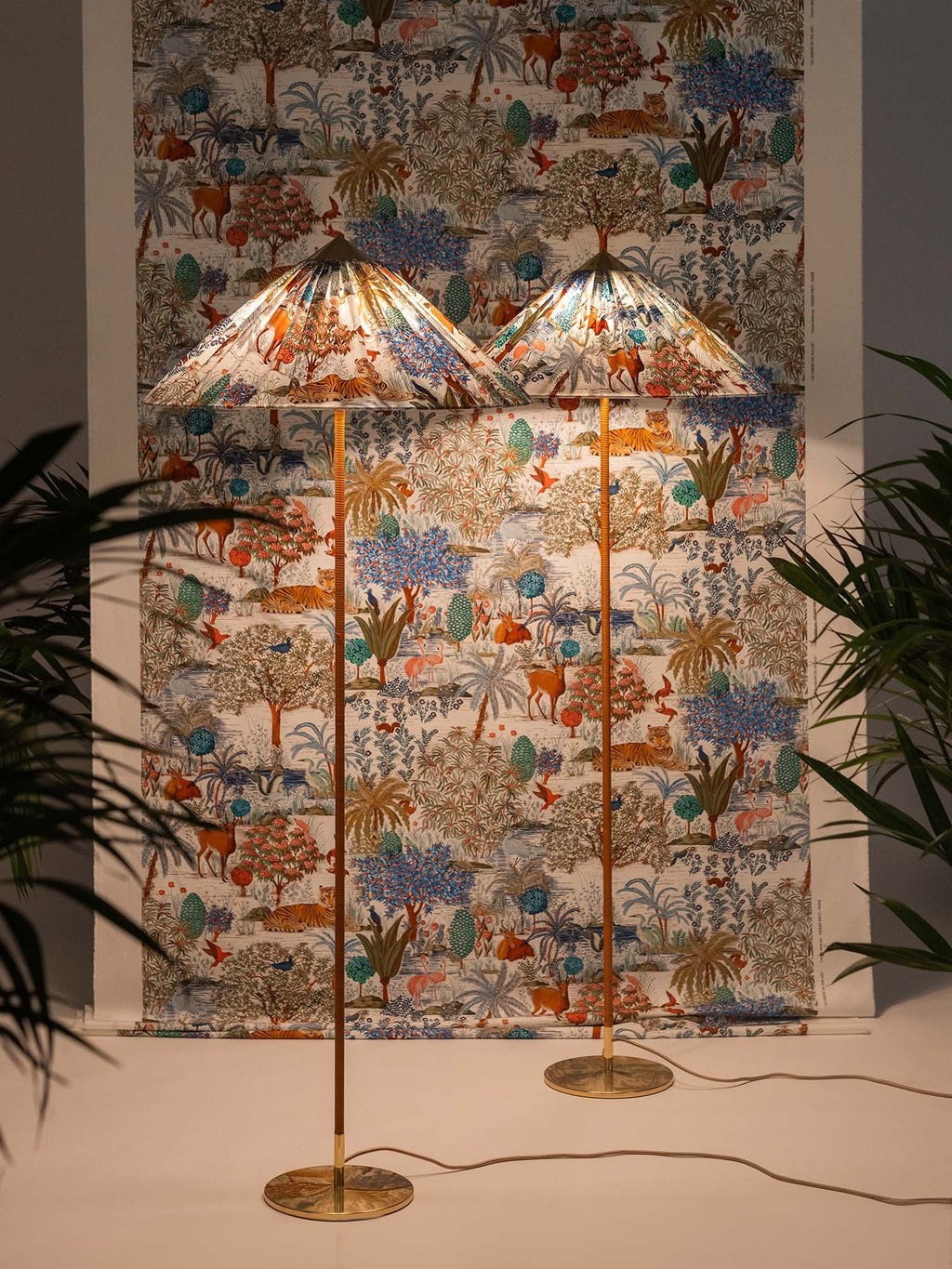 Another view of two Clérachic floor lamps with printed shades in front of a matching wall hanging, among tall plants.