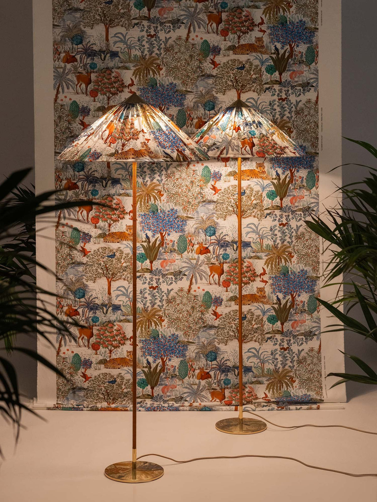 Another view of two Clérachic floor lamps with printed shades in front of a matching wall hanging, among tall plants.