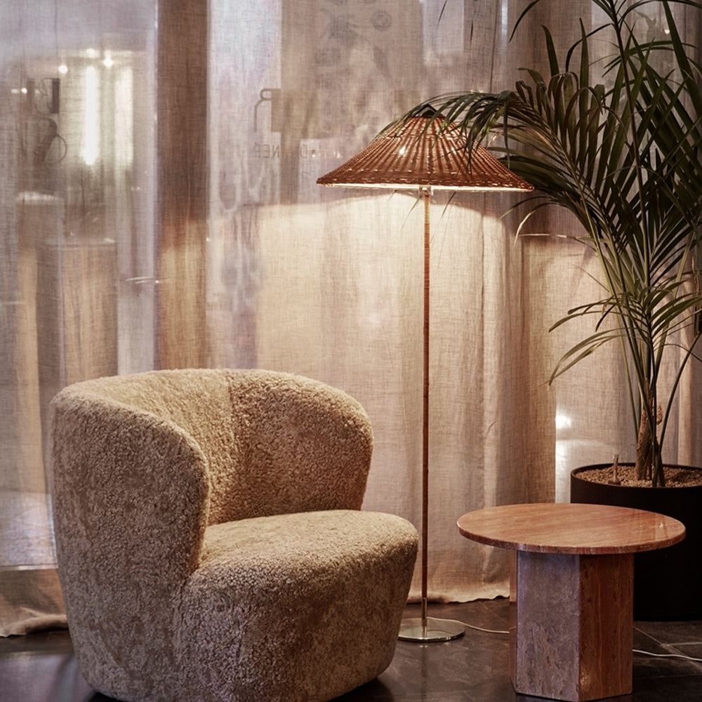 Shown here, a Clérachic floor lamp with a rattan shade glows in front of a sheer curtain, beside a shearling armchair and a stone side table.