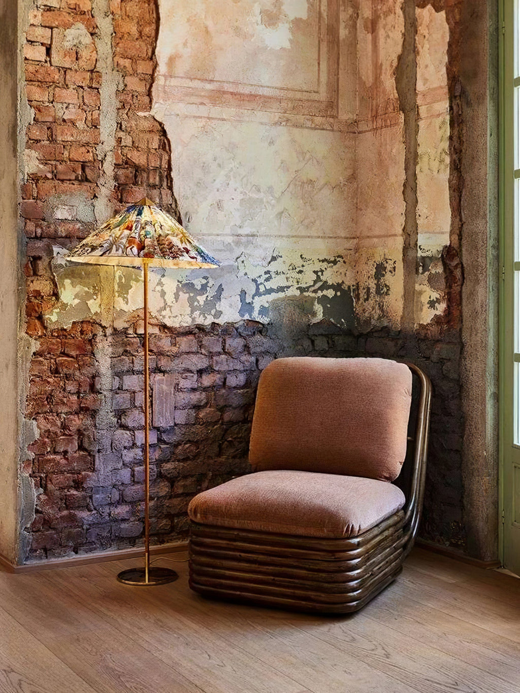 In this photo, a Clérachic floor lamp with a printed shade stands lit in a brick corner with flaking plaster, beside a low armchair in rust velvet.