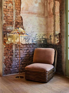 In this photo, a Clérachic floor lamp with a printed shade stands lit in a brick corner with flaking plaster, beside a low armchair in rust velvet.
