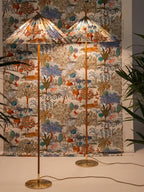 Two Clérachic floor lamps with printed shades stand in front of matching fabric, one in front of the other, among palms.