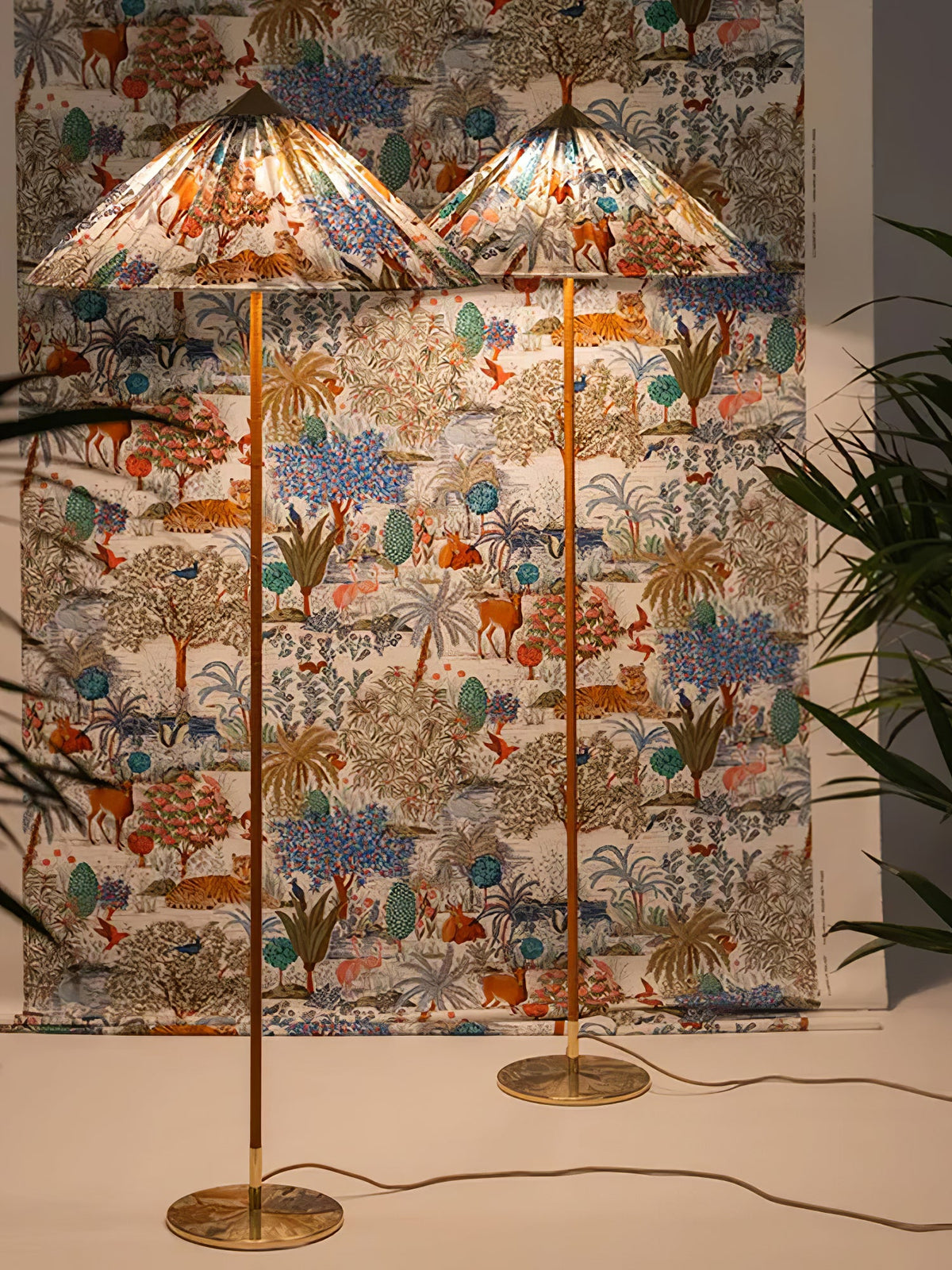 Two Clérachic floor lamps with printed shades stand in front of matching fabric, one in front of the other, among palms.