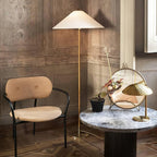 This Clérachic floor lamp with a white shade stands by a beige chair and a round marble table, with a small brass table lamp nearby.