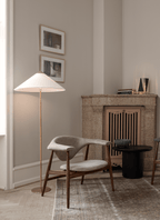 Shown here, a Clérachic floor lamp with a white shade stands in a corner beside an armchair, in a room with a stone fireplace and framed pictures.