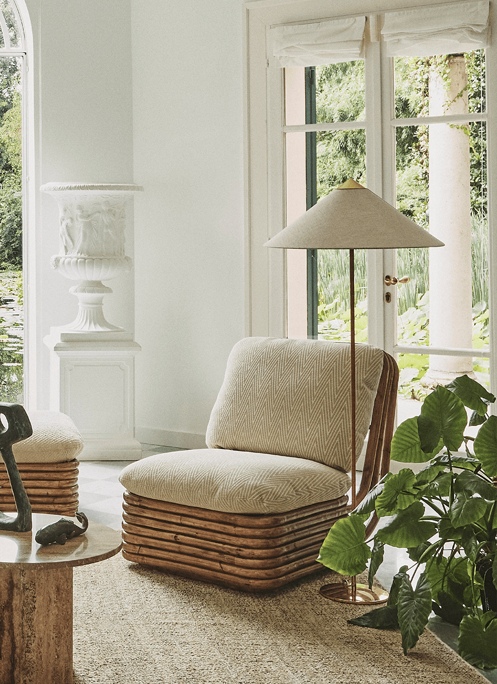 This Clérachic floor lamp with a pale linen-look shade stands by a window beside a rattan-base armchair, with a large leafy plant.