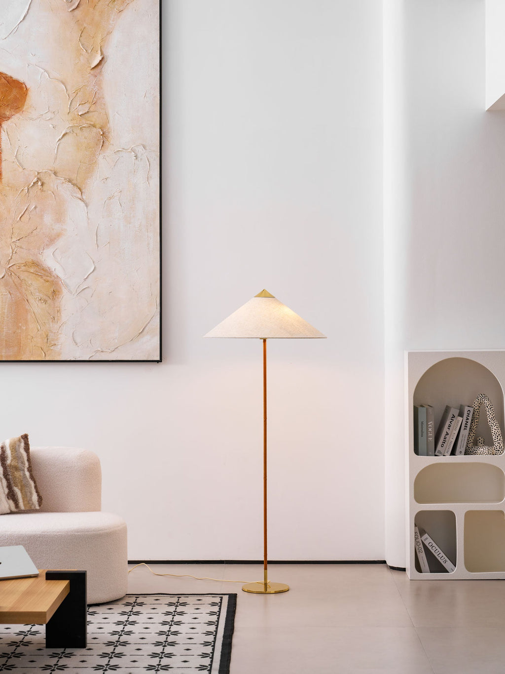 Shown here, a Clérachic floor lamp with a pale shade stands to the right of a white arched shelf with books, near a boucle chair and a patterned rug.