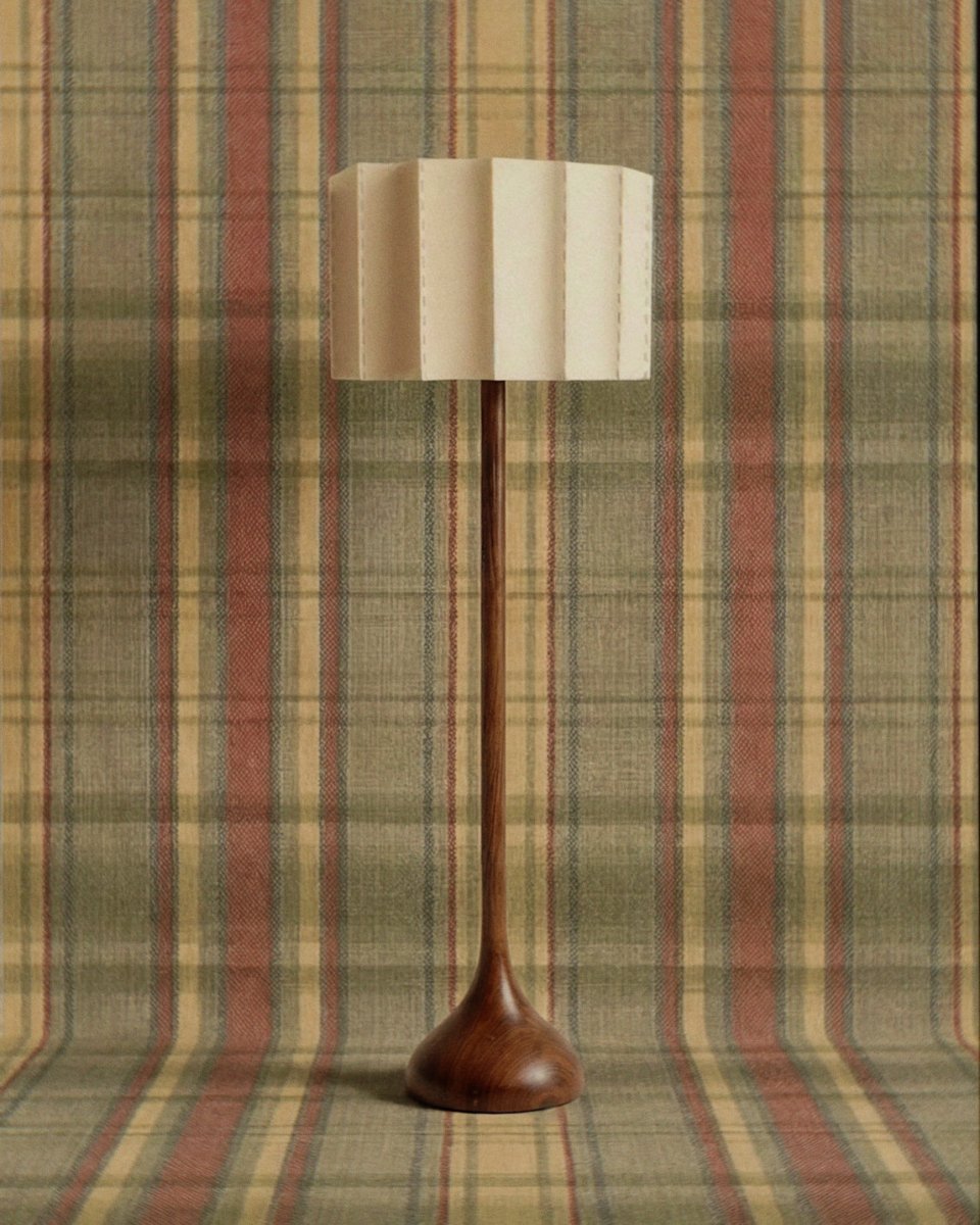 Clérachic floor lamp with a slender walnut-toned wooden stem, rounded base and pleated stitched fabric shade on a plaid backdrop.