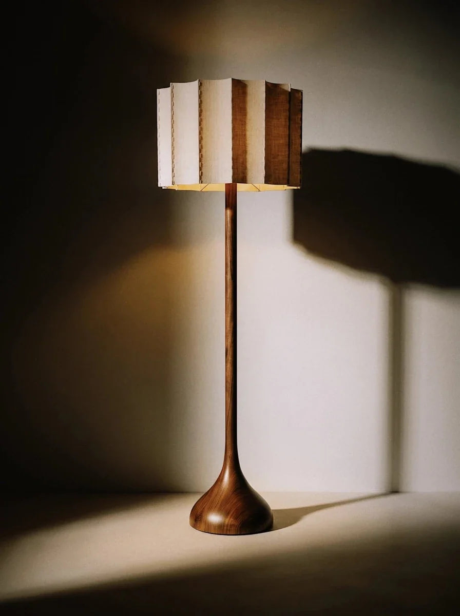 Clérachic floor lamp glowing warm against a shadowed wall, its wooden stem and pleated fabric shade casting dramatic shadows.