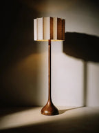 Clérachic floor lamp glowing warm against a shadowed wall, its wooden stem and pleated fabric shade casting dramatic shadows.