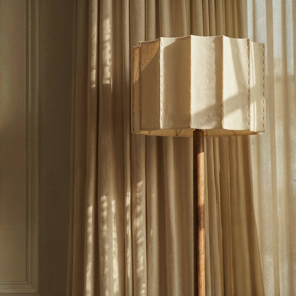 Clérachic pleated fabric shade on a wooden floor lamp, lit by afternoon sun against soft linen curtains.