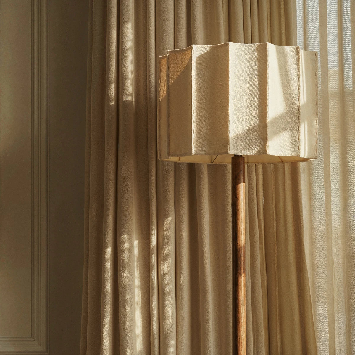 Clérachic pleated fabric shade on a wooden floor lamp, lit by afternoon sun against soft linen curtains.