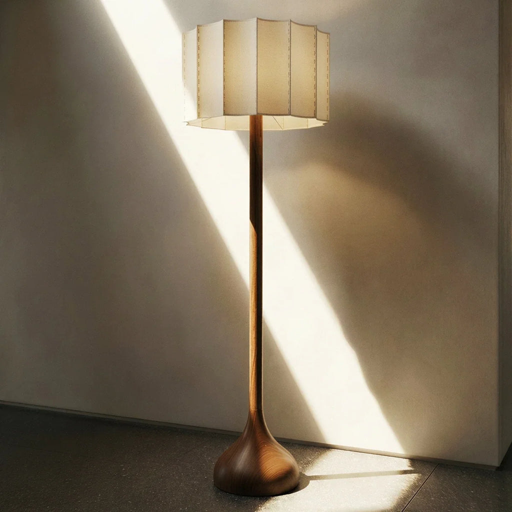 Clérachic floor lamp with wooden base and pleated shade standing in a sunbeam against a plaster wall.