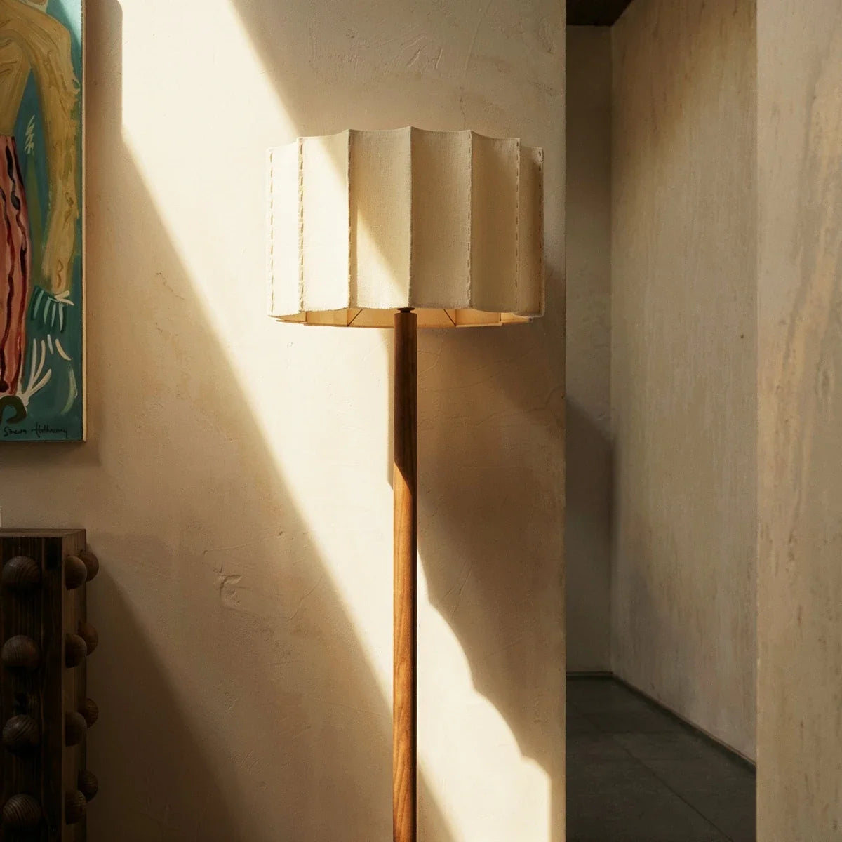 Clérachic floor lamp with pleated fabric shade and slim wooden stem in a sunlit plaster hallway beside a framed painting.
