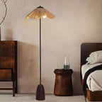 A Clérachic floor lamp with a raffia shade lights a corner with a carved wooden stool, a dark bed and a walnut cabinet.