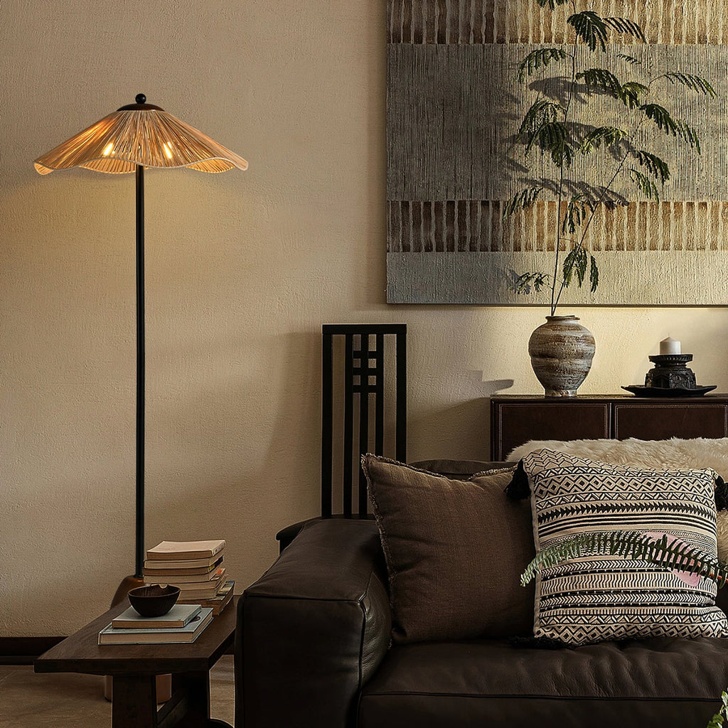 A Clérachic raffia floor lamp lit in a living room with a dark leather sofa, patterned cushions and a large textured artwork.