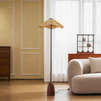 This Clérachic floor lamp's wavy raffia shade is lit by a window with long sheer curtains, beside a cream rounded armchair.
