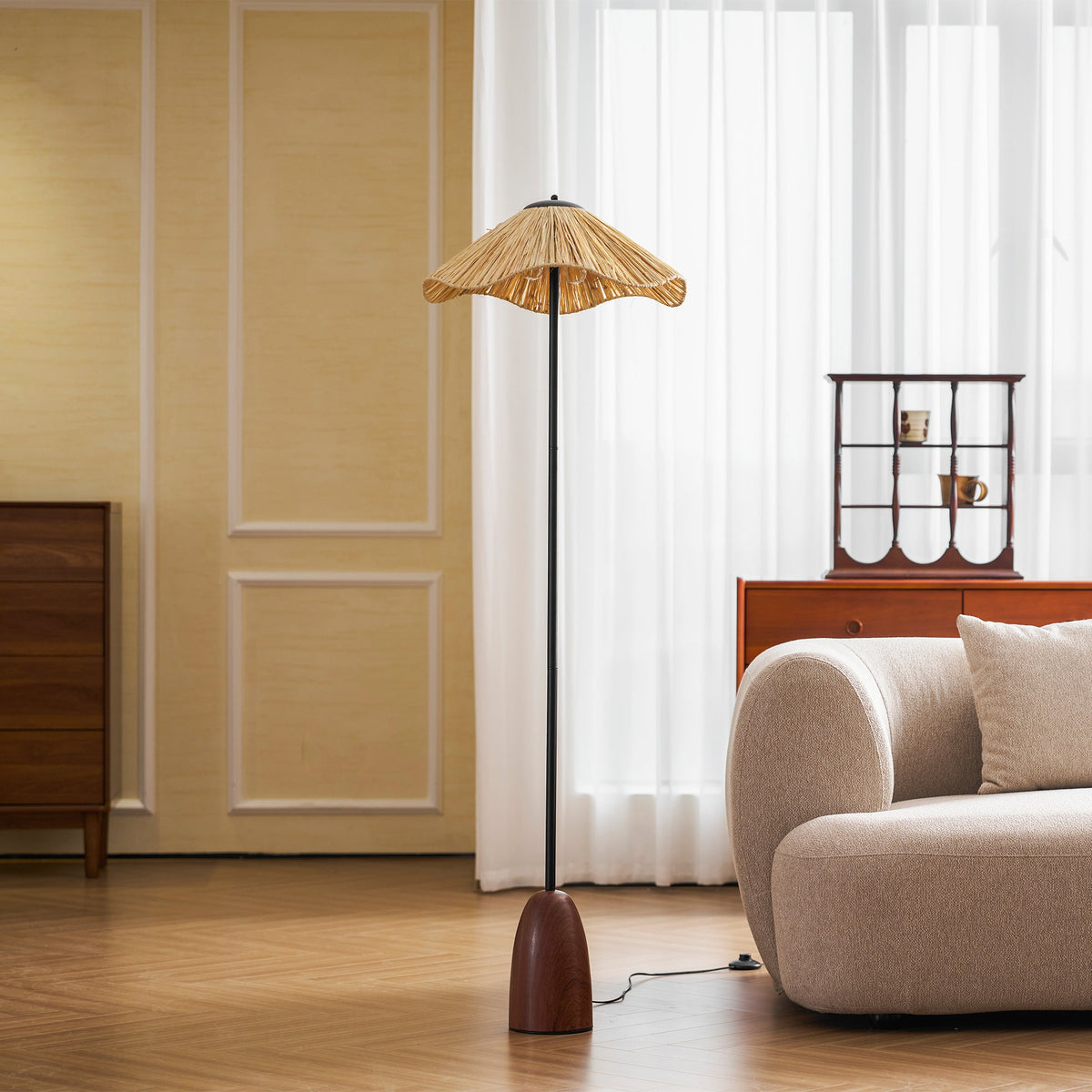 This Clérachic floor lamp's wavy raffia shade is lit by a window with long sheer curtains, beside a cream rounded armchair.