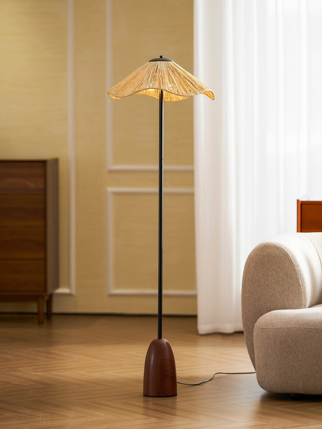 Clérachic floor lamp with a walnut base and a raffia shade, on a wooden floor beside a cream rounded sofa.