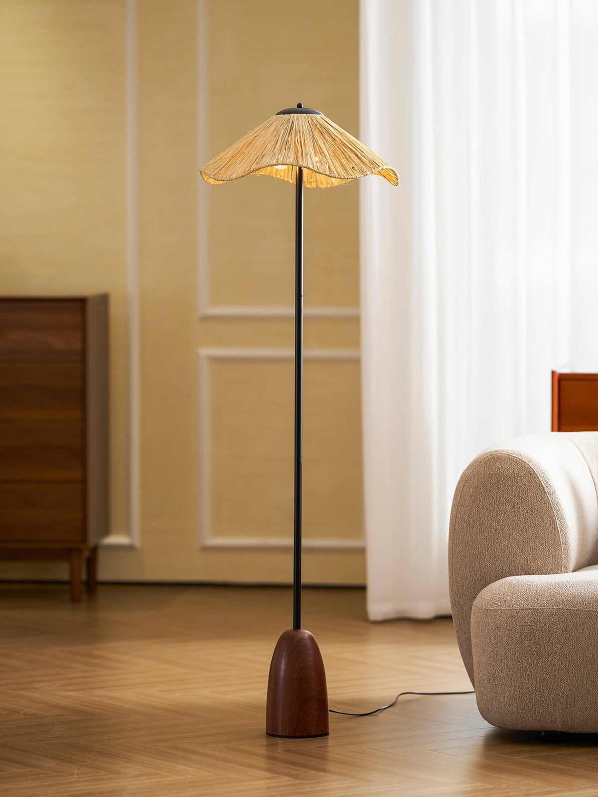Clérachic floor lamp with a walnut base and a raffia shade, on a wooden floor beside a cream rounded sofa.