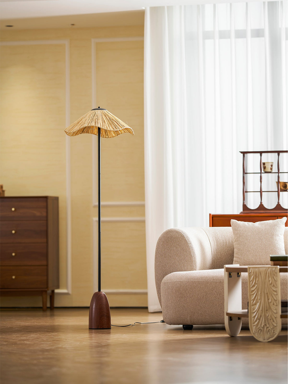 From farther back, a Clérachic raffia-shade floor lamp stands in a bright room with sheer curtains, a cream sofa and a wooden chest.