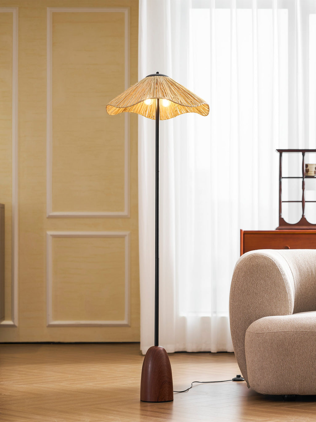 Closer view of the lit raffia shade and slim stem of this Clérachic floor lamp, with a cream sofa and sheer curtains behind.