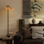 A lit Clérachic floor lamp stands beside a leather sofa with woven cushions, below a large artwork and a potted tree.