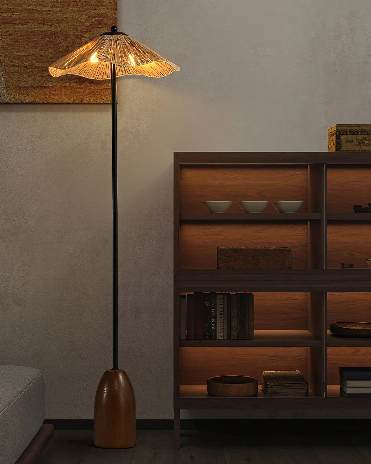 The lit raffia shade of this Clérachic floor lamp stands near a dark wooden shelving unit with bowls and books.