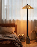 Clérachic floor lamp in a bedroom, its shade glowing behind a bed with brown bedding and a stack of books on the floor.