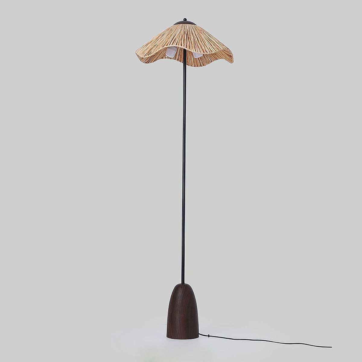 A Clérachic floor lamp with an unlit raffia shade and a walnut base, on a grey backdrop with its black cord trailing across the floor.