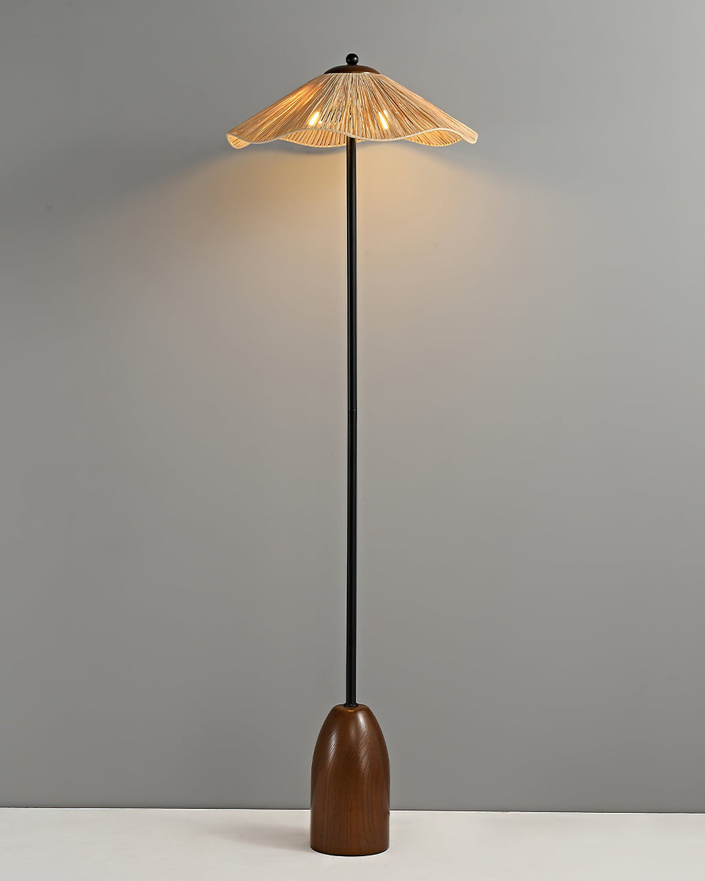 A Clérachic floor lamp with a wavy raffia shade, glowing warm, a slim black stem and a walnut base, on a grey backdrop.
