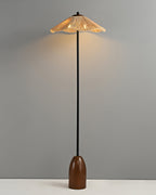 A Clérachic floor lamp with a wavy raffia shade, glowing warm, a slim black stem and a walnut base, on a grey backdrop.