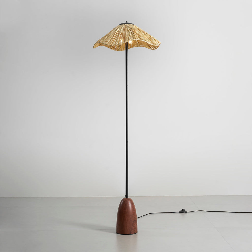 A Clérachic floor lamp with a raffia shade, a black stem and a walnut base, with an in-line foot switch on its cord, on a grey floor.