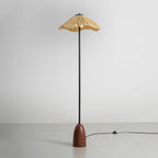 A Clérachic floor lamp with a raffia shade, a black stem and a walnut base, with an in-line foot switch on its cord, on a grey floor.