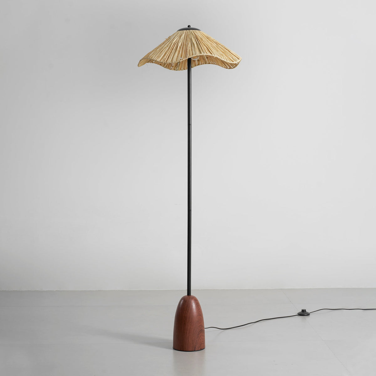 Full view of the Clérachic raffia-shade floor lamp on a grey floor, with its black cord and foot switch trailing off to the right.