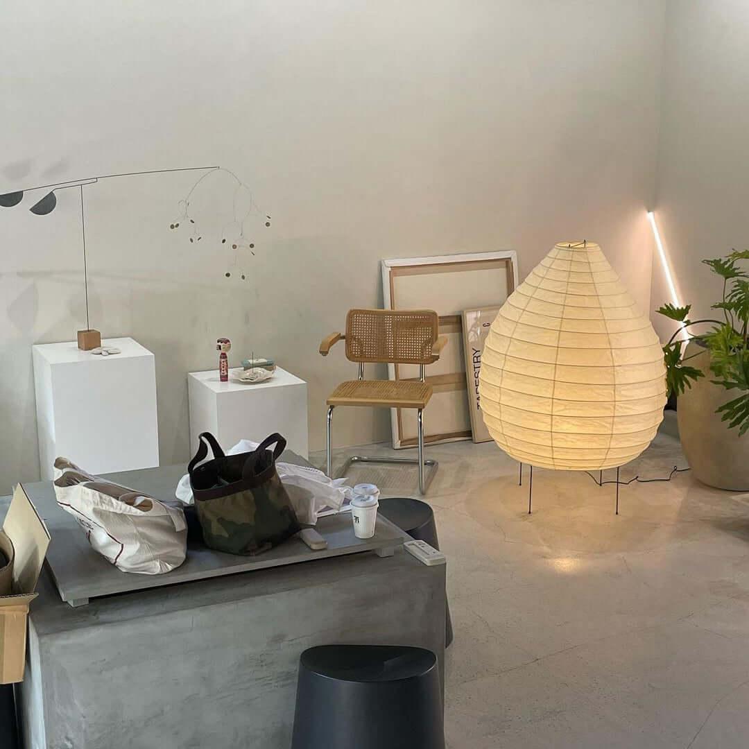Clérachic paper lantern lamp lit on a concrete showroom floor beside a cane-back chair, framed prints leaning against the wall and a potted plant.