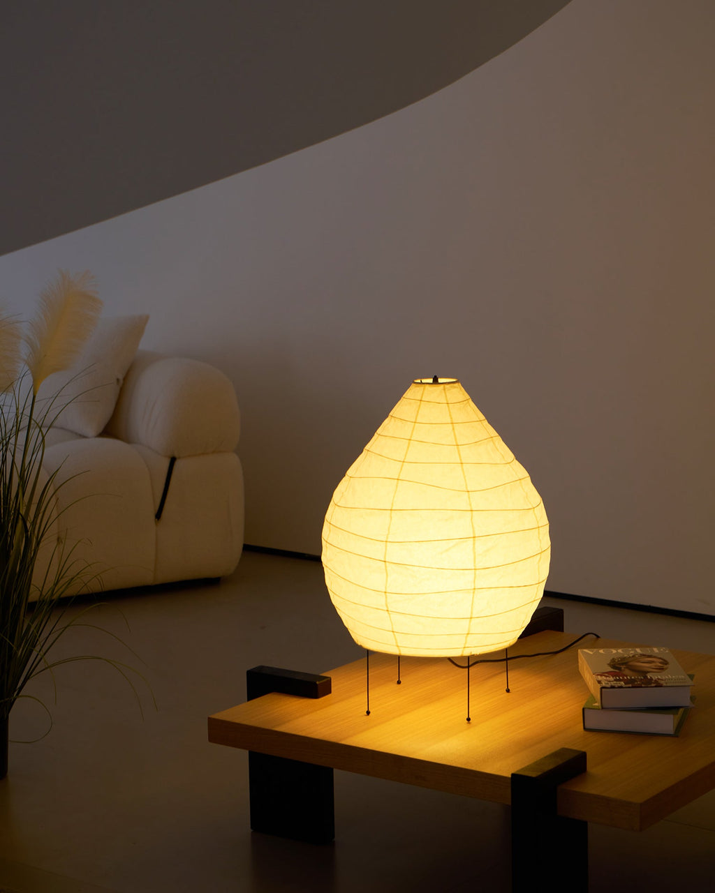 Clérachic paper lantern lamp glowing on a low wood table beside a cream bouclé sofa and a spray of pampas grass in a dim room.