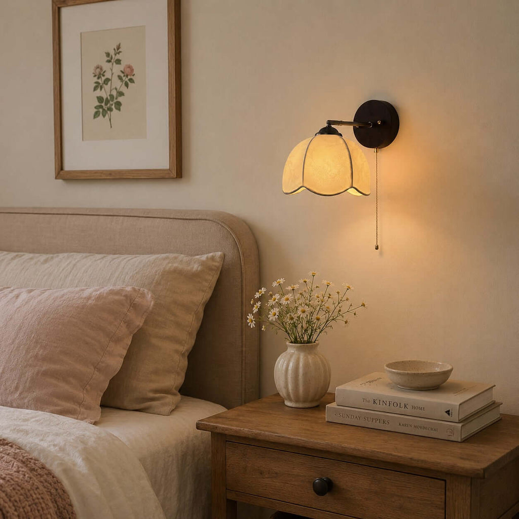 Clérachic wall light with a scalloped fabric shade glowing above a bedside table with a vase of wildflowers and stacked books.