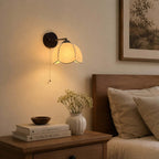 Clérachic pull-chain wall light with a dark round plate, lit above a wooden bedside table with books and a ribbed vase.