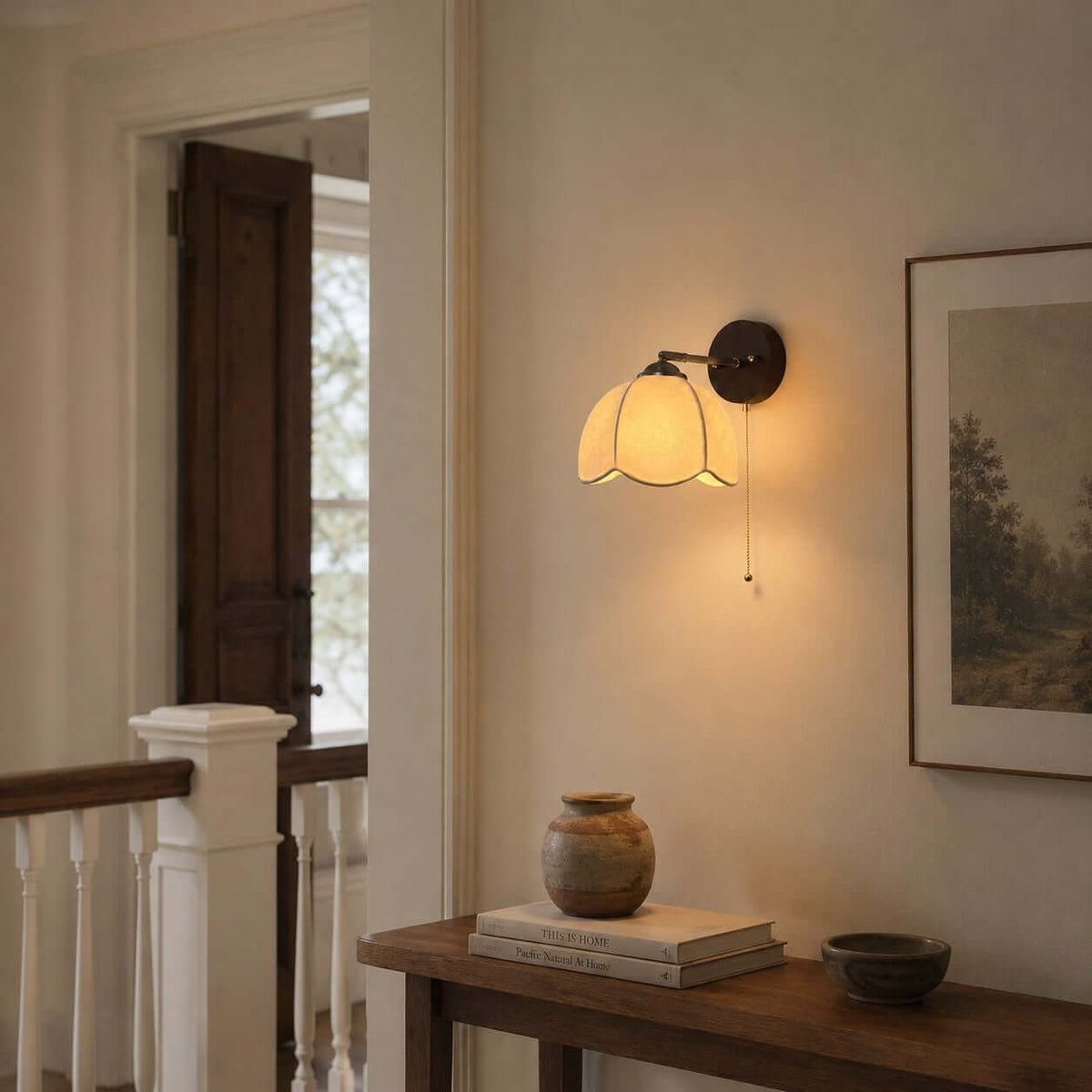 Clérachic wall light glowing beside a framed landscape at a staircase landing, above a small wooden console with a clay pot.
