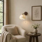 Clérachic wall light with an unlit fabric shade above a boucle armchair and a round table, with a framed botanical print.