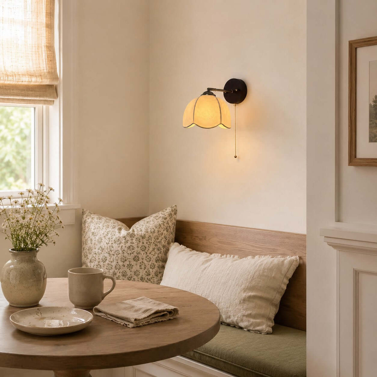 Clérachic wall light glowing above a breakfast nook with a round table, a patterned cushion and a vase of wildflowers.