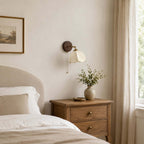 Clérachic wall light above a wooden bedside chest with a drawer pair, a cream headboard and a vase of green sprigs.