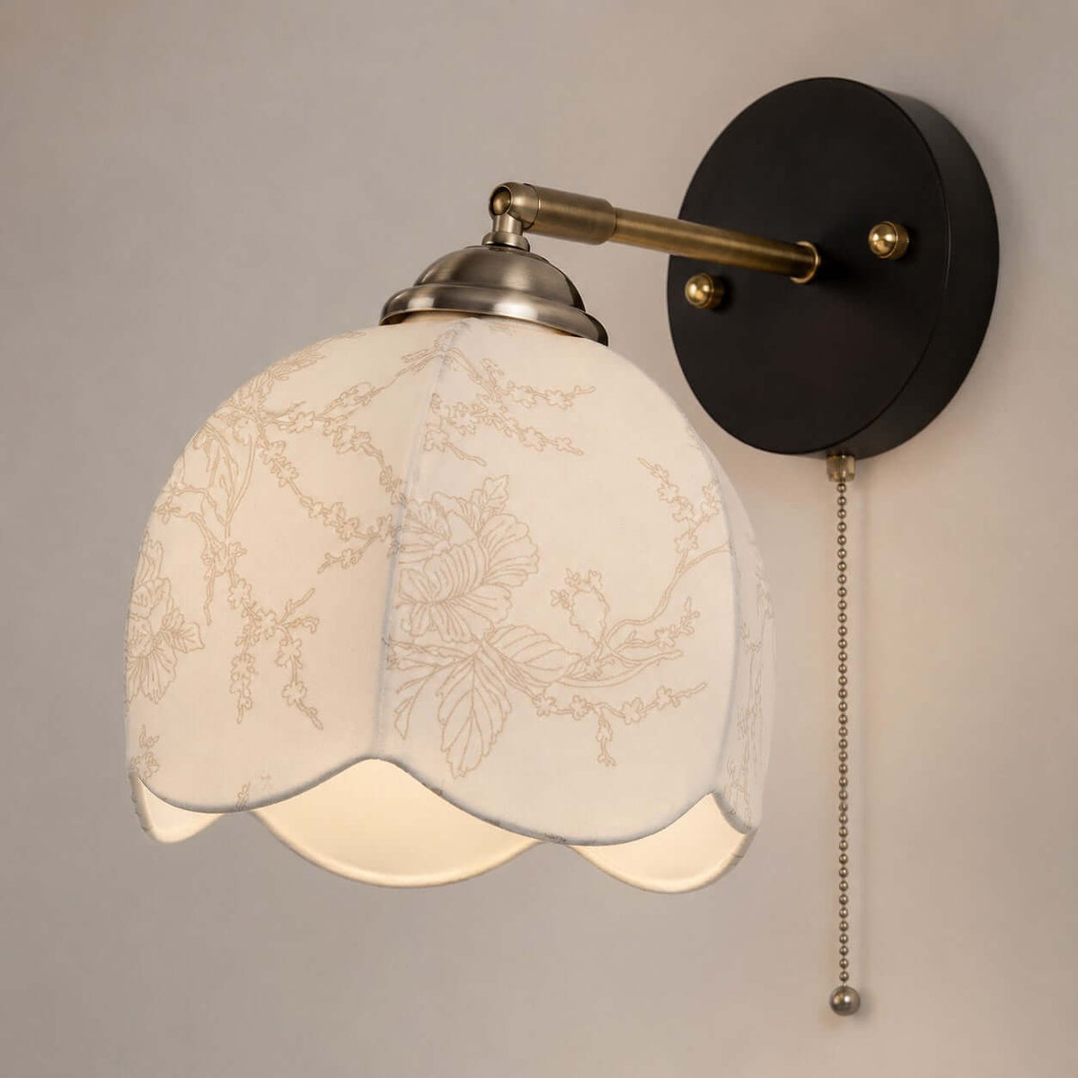 Close view of the white floral-patterned fabric shade, brass-toned arm and black plate with a pull chain on this Clérachic wall light.