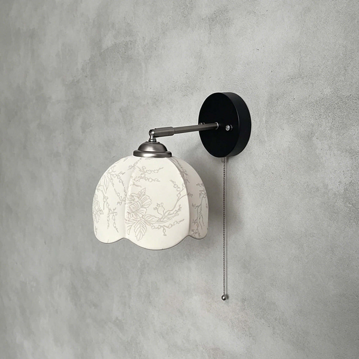 Clérachic wall light with a patterned white shade, a nickel arm and a black round plate, on a grey plaster wall.