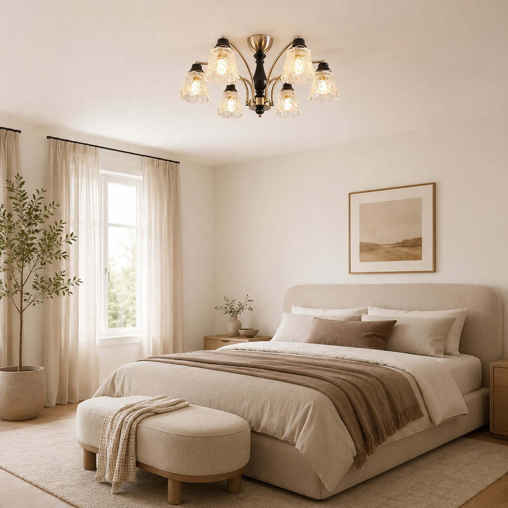 Clérachic floral-glass chandelier over a bed with a rust and taupe throw and a round upholstered bench, with sheer curtains beside.
